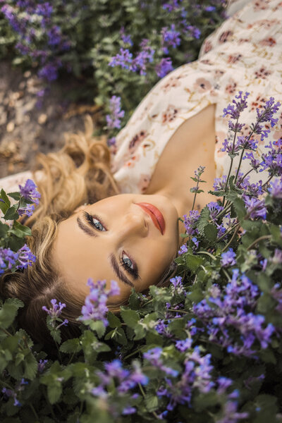senior girl with blonde hair looking at the camera and laying in a patch of small purple flowers