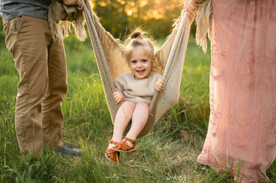 Little girl sitting in a blanket swing, smiling at the camera in a filed in Ottawa