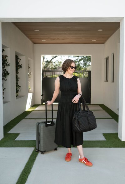 The owner of Scout & Bimble stands under a covered outdoor walkway with her suitcase and travel bag, wearing a black dress and orange sneakers.