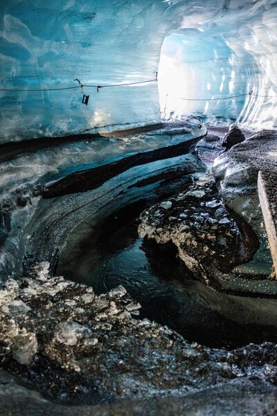 A bride and groom look at each other smiling and laughing happily after popping a bottle of champagne after their elopement. They are standing at the bottom of a canyon surrounded by red dirt, green trees and white and orange canyon walls. The bride is in a flowy white dress and the groom is in a navy blue suit.