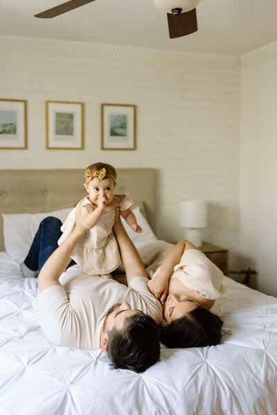 Young parents lifting their toddler into the air while lying on their bed during a family photoshoot.