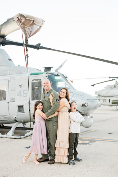 A pilot father stands hugging his wife and young daughter in front of a military helicopter with son leaning on mom