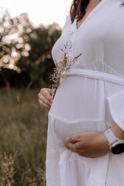 Eine schwangere Frau steht auf einer Wiese in einem weißen Kleid und hält ihren Babybauch