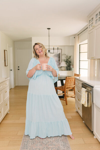 Mollie Mason in a long blue dress holding a coffee mug and standing in the middle of her kitchen