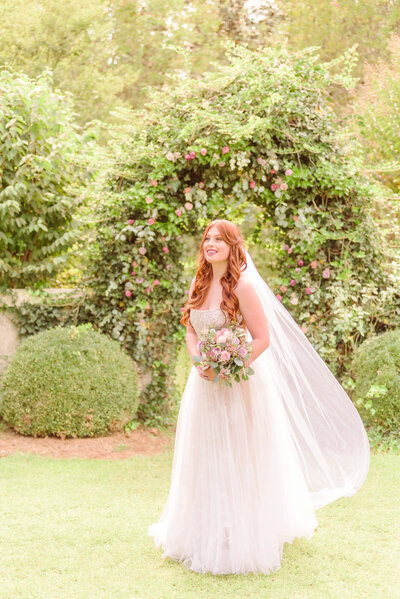 Olivia smiles to the side, there is a green archway adorned with roses behind her.