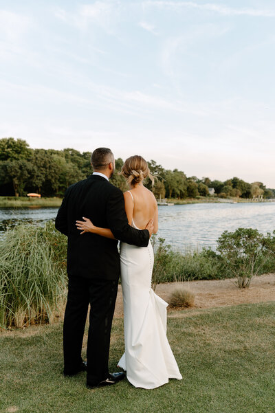 A newlywed couple stands arm-in-arm by the tranquil waterfront at Old Field Club, gazing out at the peaceful bay as the sun begins to set. The bride, dressed in an elegant backless white gown with delicate buttons trailing down the train, rests her hand gently on her groom’s back. Her hair is styled in a soft, romantic updo. The groom, wearing a classic black tuxedo, holds his bride close as they take in the serene moment together. Lush greenery and tall grasses frame the shoreline, while distant docks and waterfront homes add to the idyllic coastal charm of this Long Island wedding venue. The sky is painted in soft pastel hues, enhancing the dreamy, romantic atmosphere of their special day.