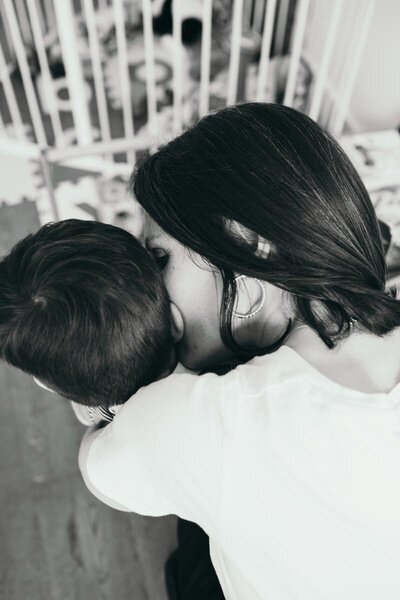 Black-and-white photo of Kaya hugging her young child Luca close, facing away from the camera.