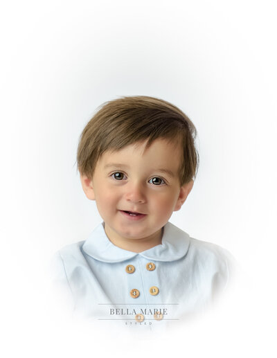 One year old little boy with dark brown hair and big brown eyes, smiling with a blue shirt with brown buttons, white vignette background.
