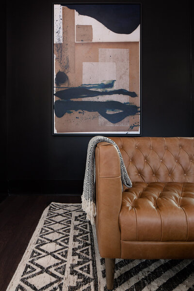 Black wall with painting. Brown leather couch and rug in forground
