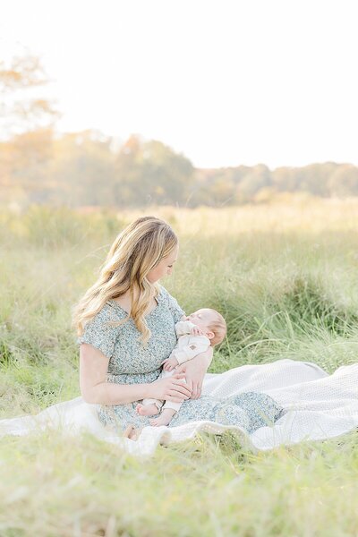 mom sits in field with baby during outdoor newborn photo session at Heard Farm in Wayland Massachusetts with Sara Sniderman Photography