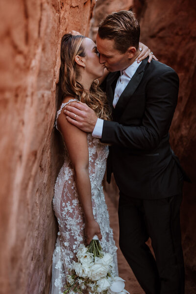 bride and groom kissing while leaning against rock wall in the desert