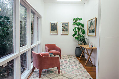 The image features a cozy interior space with a large window on the left allowing natural light and greenery views. Two plush, coral-colored armchairs with a matching cushion on one face inward, positioned on a multicolored patterned area rug. The right side of the room shows a small wooden desk with a white vase containing pink flowers and a potted green plant. Above the desk, there are three framed pictures on the wall, each depicting floral or botanical scenes. The room has white walls and a light wooden floor, creating a bright and welcoming atmosphere.