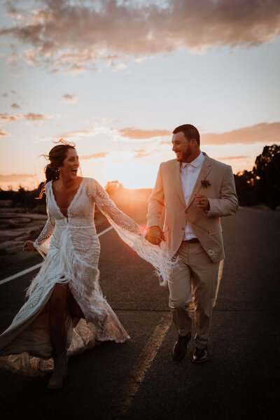 romantic bride and groom holding hands and running away from a beautiful sunset at dead horse state park