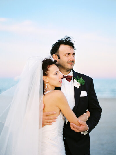 Bride and groom embrace, looking off into the distance on the beach