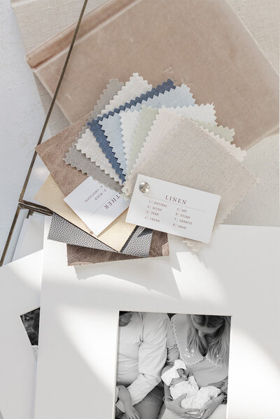 Two matted prints stacked on top of a glass print box with an heirloom album and fabric swatches. The matted print on top shows a picture of a mother holding a baby with the father next to her by Katie Stansfield Photography, a family photographer in Richmond.