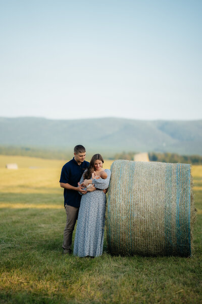man and woman standing next to a bale of hay while holding their newborn baby