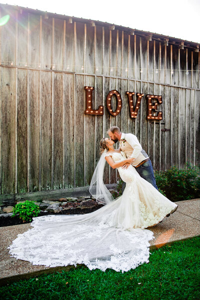 Groom dipping bride for a dramatic kiss next to the Love letter sign on the side of the barn