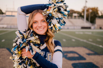 High school cheerleader poses for the camera with pom poms on the football field.