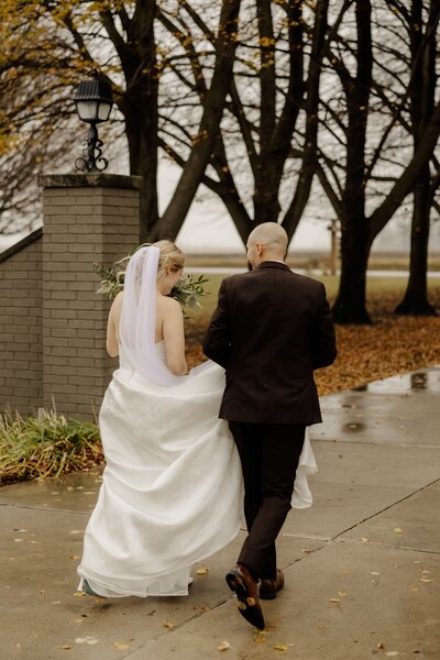 A bride and groom run through the rain together as groom hold's her dress.