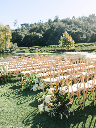 Rows of elegant wooden chairs with white cushions are set on lush green grass, surrounded by vibrant floral arrangements, against a serene forest backdrop.