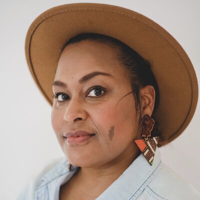 Afro-Latina woman wearing a tan hat, triangular earings with a black power fist, and a light blue chambray shirt
