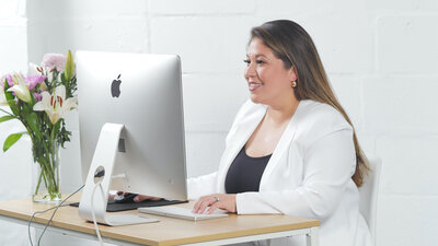 Teresa sits at a desk while using a Mac computer. There is a bouquet sitting on the desk as well with an assortment of pink, purple, and white flowers.