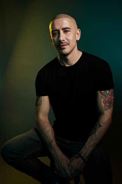 Moody portrait of Ray Contreras in a dark setting, wearing a black t-shirt and seated confidently, captured by Ylva Erevall.