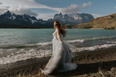 A lesbian couple in white wedding attire pop champagne. They are both smiling and laughing, the champagne is spraying and the mountain peaks of Triglav National Park in Slovenia are in the background.