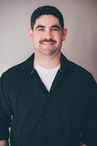 A man with short dark hair and a mustache, wearing a black button-up shirt over a white T-shirt, stands against a plain background, smiling as if pleased with his latest web design and branding project.