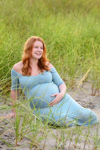 pregnant lady with red hair wearing a blue dress sitting on the beach for her outdoor maternity photos.