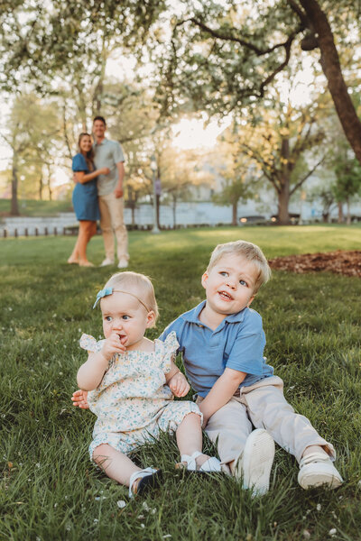 Toddler brother sits with his little sister as mom and dad look adoringly in the background