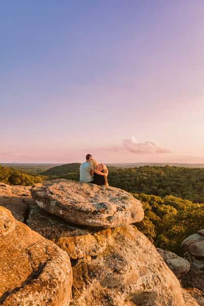 couple sitting on a rocky overlook at Garden of the Gods in Southern Illinois during their engagement session, bathed in golden hour light with a panoramic view of the forest below.