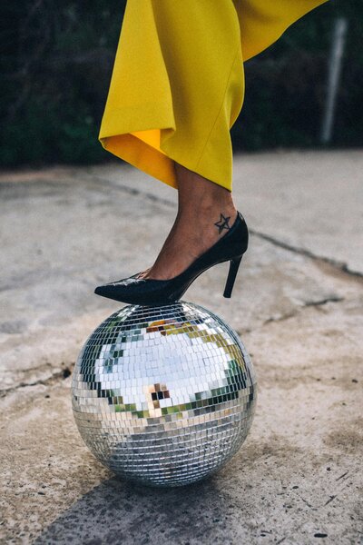 Woman in yellow pants holds down a disco ball with her black heel covered foot