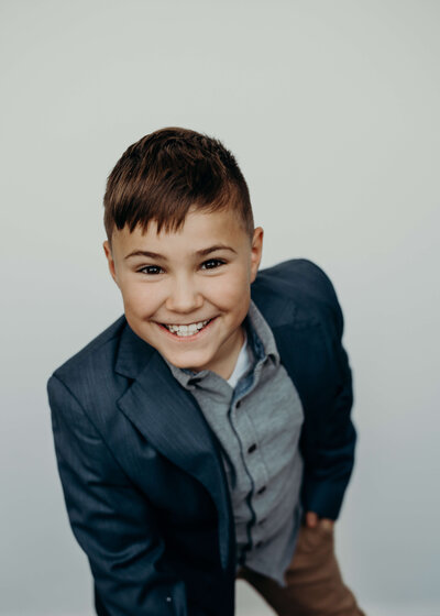 An eight year old boy grins playfully at the camera as he leans forward towards the audience, wearing a navy suit coat what happened and button up shirt.