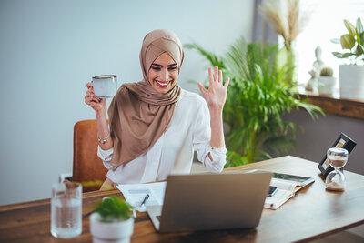 A Muslim woman with a headscarf smiles and holds her hands up while on a video call. She has a coffee cup in one hand