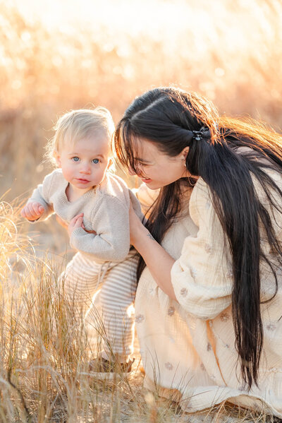 A mom holds onto her toddler son while he looks at the camera during a family session with Justine Renee Photography. Both mom and son wear neutrals, complimented by the tall brown grass around them.