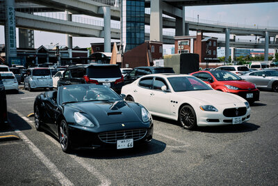 A luxury car lineup featuring a black Ferrari California and white Maserati Quattroporte parked at Daikoku PA in Tokyo, ready for a private guided tour with Tokyo Luxe Drive.
