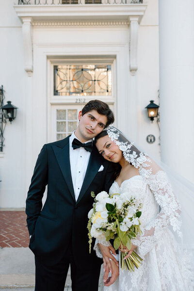 Bride leaning on groom’s shoulder with bouquet in front of white building