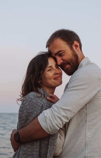 A man hugging his wife while both are smiling. This can represent the relief couples in the aftermath of infidelity can feel after affair recovery.You can benefit from affair recovery program by Relationship Experts in the United States, Canada, the UK, and worldwide. Schedule a free consultation to start healing.