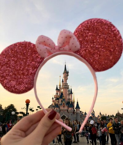 Hand holding pink sparkly Minnie Mouse ears up so the castle at sunset is visible through the headband’s circular opening, city crowds blurred behind.
