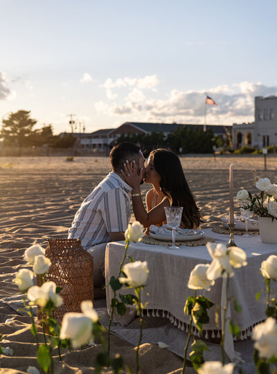 Couple sharing a kiss during a romantic beachside dinner setup with white roses and elegant table décor, styled by Yours Truly, a wedding planner and designer in New Jersey.
