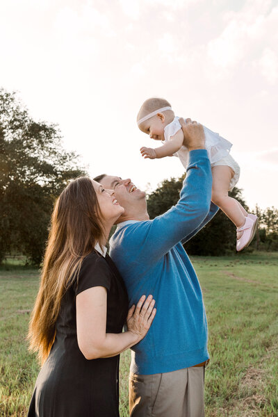 Smiling parents enjoying a sunny outdoor moment with their baby, as the father lifts the baby girl into the air while the mother lovingly watches. A joyful family photography session in a grassy field.