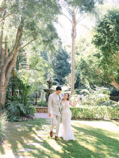 A couple at Hartley Botanica Garden in wedding attire walks happily through a sunlit garden surrounded by lush greenery.