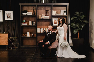 A bride in a sleek white gown stands beside her seated groom in a modern lounge space at a Chandler wedding and event venue. The couple poses in front of a stylish wood bookshelf with curated décor, combining elegance with a contemporary indoor setting.