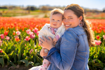 Smiling mother in a denim jacket holding her baby among vibrant tulips during a spring photo session – captured by Capture Joy Photography, Northern Virginia family photographer