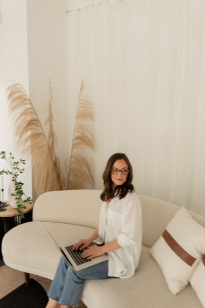 Female SEO Consultant sitting on sofa with laptop wearing jeans and white linen button up looking over her shoulder