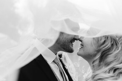 bride and groom touching noses underneath a white vail and smiling