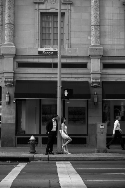 A couple leans against a streetlight on a city sidewalk, with historic architecture and pedestrians in black and white surroundings.