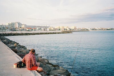 Man in a red plaid shirt sits on a rock-lined jetty with a fishing pole, facing a quiet coastal town at dusk.