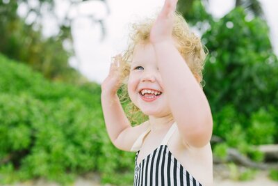 A little girl with curly blonde hair, hold her hands up in the air, smiling.